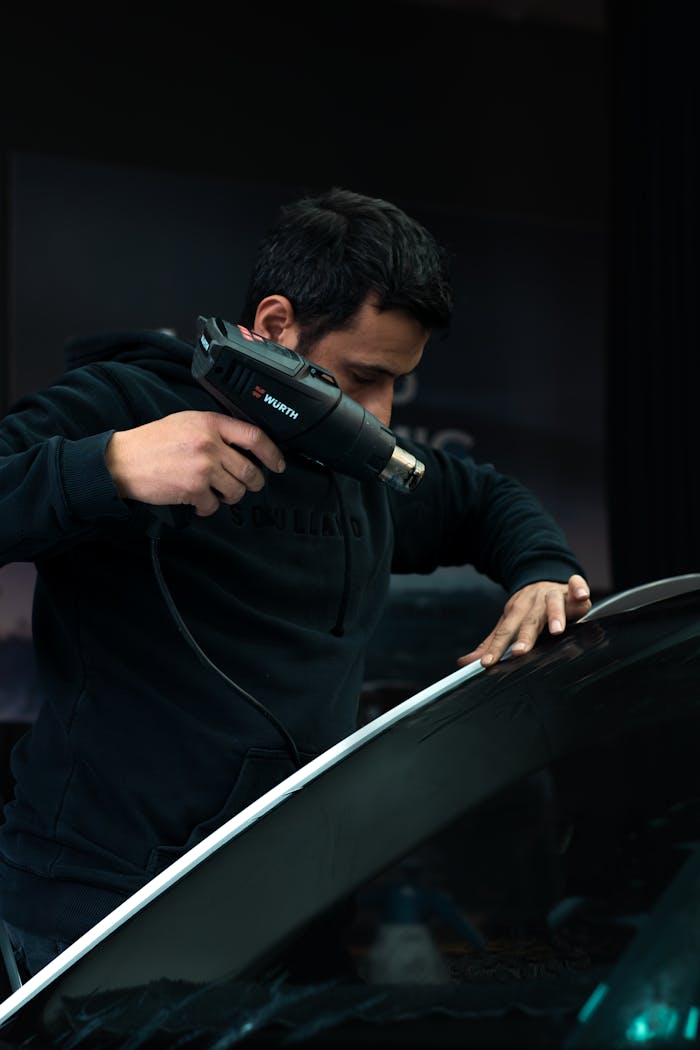 About Mechanic carefully applies heat to car windshield with a heat gun in a garage setting.